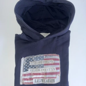 Mörkblå hoodie från Ralph Lauren - Säljer en snygg mörkblå hoodie från Ralph Lauren med en stor patch framtill som har ett amerikanskt flaggmönster och text. Perfekt för en avslappnad stil. Storlek M. Inga skador mm