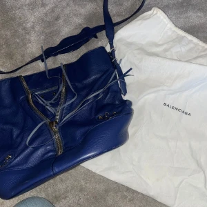 Mörkblå axelväska från Balenciaga - Säljer en snygg mörkblå axelväska från Balenciaga i äkta skinn. Väskan har dragkedja framtill, justerbar axelrem och flera små fack med dragkedjor. Perfekt för dig som vill ha en stilren och lyxig accessoar.