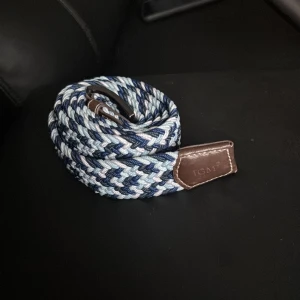 Blått flätat bälte med läderdetaljer - Säljer ett snyggt flätat bälte i olika blå nyanser med bruna läderdetaljer från IGAN. Bältet har ett randigt mönster och passar perfekt till jeans eller chinos. Spännet är i metall och bältet är flexibelt och bekvämt.