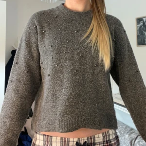 Grå stickad tröja med pärlor - Mysig grå stickad tröja med pärldetaljer framtill. Tröjan har rund halsringning, långa ärmar och en något croppad passform. Perfekt för kyliga dagar när du vill ha något både snyggt och bekvämt.