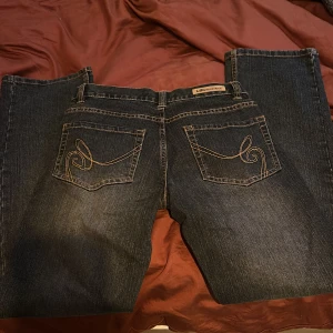 Mörkblå LÅGMIDJADE jeans - Mörkblå jeans med raka ben och klassiska kontrastsömmar. Snygga broderade detaljer på bakfickornal. Midja är 68 cm och innerbenslängd är 73 cm.
