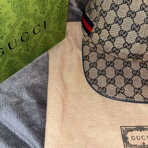 Beige keps från Gucci med monogram - Snygg beige keps från Gucci med det klassiska GG-monogrammet. Kepsen har en justerbar rem baktill och en röd och blå randdetalj på sidan. Perfekt för att ge din outfit en lyxig touch. Kan ta emot byten!! 