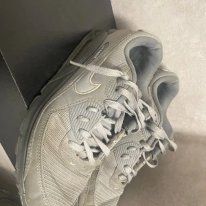 Grå Nike Air Max sneakers - Säljer ett par grå Nike Air Max sneakers med klassisk design och synlig luftdämpning i sulan. Skorna har snörning och är tillverkade i syntetmaterial med detaljer i olika grå nyanser. Perfekta för en sportig look.