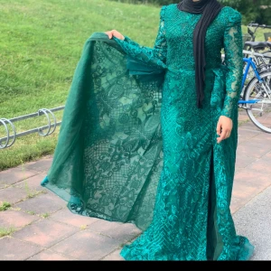 Grön långklänning med spetsdetaljer - Elegant grön långklänning med vackra spetsdetaljer och pärlor. Klänningen har långa ärmar och en slits framtill. Perfekt för en festlig tillställning. Storlek 38-40 och köpt för 5000 kr 