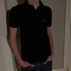 Svart pikétröja från Lacoste - Säljer en klassisk svart pikétröja från Lacoste med den ikoniska krokodilloggan på bröstet. Tröjan har korta ärmar och en stilren krage med knappar. Modellen är 176cm lång.