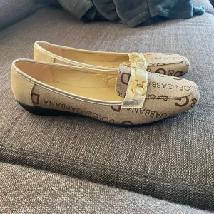Loafers från Dolce & Gabbana - Snygga beige loafers från Dolce & Gabbana med logotypmönster och gulddetaljer. Skorna har en elegant design med en metallspänne framtill och är perfekta för en stilren look. ”Hålet” som är i sulan är den enda skavanken 