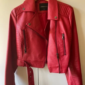 Röd skinnjacka från Sally and Dog - Snygg röd skinnjacka från Sally and Dog i storlek XL men är mindre i storlek och passar till folk som bär M i storlek. Jackan har en cool bikerstil med dragkedjor och ett bälte i midjan. Perfekt för att ge din outfit en edgy touch. 🖤