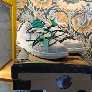Nike Dunk Low Off White - Säljer ett par Nike Dunk Low Off White sneakers i vitt och grått med gröna och svarta detaljer. Skorna har en unik design med extra snörning och en färgglad etikett på sidan. Perfekta för sommaren. Jag är öppen för frågor eller förslag!