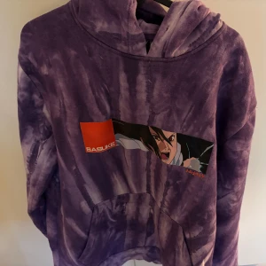 Lila hoodie med anime-tryck från Cropp - Säljer en cool lila hoodie från Cropp med ett anime-tryck på framsidan. Hoodien har en tie-dye effekt och en stor ficka framtill. Perfekt för dig som älskar anime och vill ha en bekväm och stilren tröja.