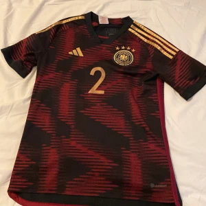Tyskland fotbollströja från Adidas - Snygg Tyskland fotbollströja från Adidas med guldiga detaljer och nummer 2. Tröjan har korta ärmar och ett coolt mönster. Perfekt för fotbollsfans!