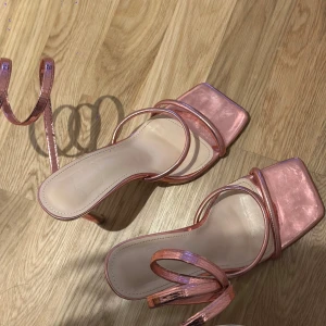 Rosa sandaler med remmar och klack - Snygga rosa sandaler med glittrig finish och smala remmar som snörs runt ankeln. De har en fyrkantig tå och en liten klack, vilket ger en elegant look. Perfekta för att sätta en glamorös touch på din outfit. Endast använt en gång!