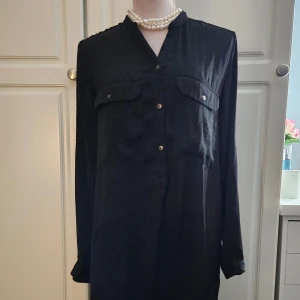 Svart klänning/tunika nyskick - Svart klänning/tunika med långa ärmar och två bröstfickor. Klänningen har en avslappnad passform och knäpps med knappar framtill. Perfekt för både vardag och fest. Den har en knapp på armarna så man kan vika upp och få trekvartsärm. Liten slits på sidorna, längre baksida. Stor I Storleken.