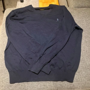 Mörkblå tröja från Polo Ralph Lauren - Säljer en stilren mörkblå tröja från Polo Ralph Lauren med broderad logotyp på bröstet. Tröjan har en klassisk v-ringning och långa ärmar, perfekt för en avslappnad men elegant look.