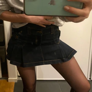 Mörkblå jeanskjol med knappar - Snygg mörkblå jeanskjol med knappar och dekorativa sömmar. Kjolen har en kort längd och en klassisk design med fickor och bälteshällor. Perfekt för en trendig look!