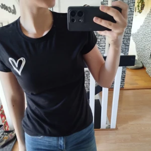 Svart t-shirt med hjärtdetalj - Säljer en svart t-shirt med ett vitt hjärta tryckt på bröstet. T-shirten har en normal passform och korta ärmar. Perfekt för en avslappnad stil.