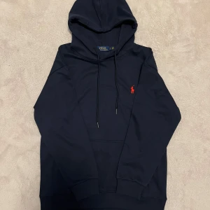 Mörkblå hoodie från Ralph Lauren - Säljer en stilren mörkblå hoodie från Ralph Lauren med det klassiska röda logotypbroderiet på bröstet. Hoodien har en justerbar huva med snören och en stor magficka framtill. Perfekt nu till våren för att fräscha upp sin stil ännu mer! 