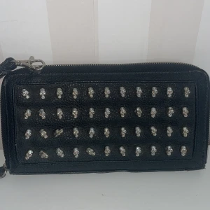 Svart väska/plånbok med dödskallar från Gina Tricot - Cool svart plånbok från Gina Tricot dekorerad med små silverfärgade dödskallar. Plånboken har en dragkedja och flera fack inuti för kort och kontanter. Perfekt för att ge din stil en edgy touch! Mått 23*12 cm.