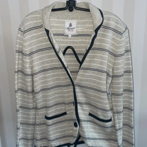 Gråblå randig kofta från Honey ny - Säljer en stilren beige kofta från Honey med svarta ränder och knappar. Koftan är tillverkad i 100% bomull och har en klassisk design med fickor framtill. Perfekt för en avslappnad look.