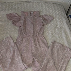 Glittrig rosa jumpsuit från Lindex - Säljer en superfin glittrig rosa jumpsuit från Lindex för barn. Den har korta ärmar med volangdetaljer och en bekväm passform. Perfekt för festliga tillfällen! ✨