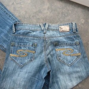 Blå jeans med broderade bakfickor - Snygga blå jeans med unika broderade detaljer på bakfickorna. De är bootcut-stil och nsidan av midjan har ett färgglatt mönster som ger en extra touch. Midjemått: 36cm Grentmått: 17cm Innerbenslängd:84cm