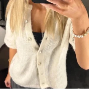 Vit ribbad kofta med pärlknappar - Säljer en stilren vit ribbad kofta med korta ärmar och vackra pärlknappar. Perfekt för en elegant look. Passar bra till både jeans och kjol.