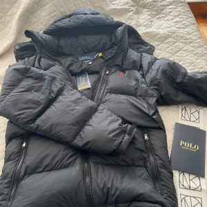 Svart dunjacka från Polo Ralph Lauren - Säljer en svart dunjacka från Polo Ralph Lauren. Jackan har en praktisk dragkedja och är långärmad med en mysig huva. Skriv vid minsta funderingar, pris kan diskuterar inga skador på jakan säljer den då den är för stor och dags för en ny jacka. 