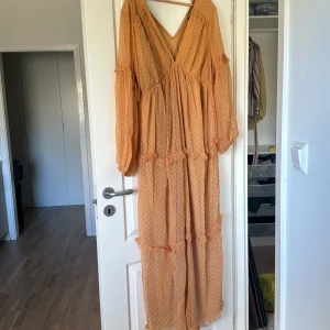 Beige långklänning med volangdetaljer - Säljer en elegant beige långklänning med v-ringning och volangdetaljer. Klänningen har långa ärmar och ett diskret prickigt mönster. Perfekt för en stilfull look.
