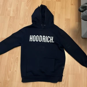 Mörkblå hoodie från Hoodrich - Säljer en svart hoodie från Hoodrich med vit logga på bröstet. Den har en klassisk känguruficka och justerbar huva med snören. Perfekt för en avslappnad stil.