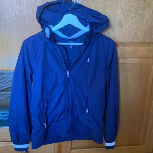 Blå vindjacka från Polo Ralph Lauren - Säljer en snygg blå vindjacka från Polo Ralph Lauren. Jackan har en praktisk huva och dragkedja framtill. Den är perfekt för blåsiga dagar och har ribbade muddar vid ärmsluten. En klassisk logga pryder bröstet.