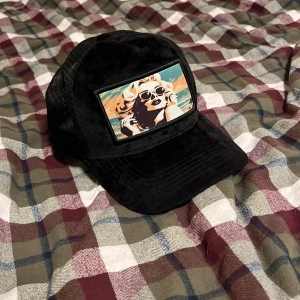 Svart keps med tryck från Revenite - Snygg svart keps från revenite med ett coolt tryck av en kvinna i solglasögon på framsidan. Kepsen har ett nätmaterial baktill för bra ventilation och justerbar passform. Perfekt för en avslappnad stil.(helt ny)