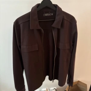 Mörkbrun jacka med dragkedja - Säljer en stilren mörkbrun overshirt från dressmann med dragkedja framtill. Jackan har en klassisk krage och långa ärmar, perfekt för en elegant look. Passar bra till både vardag och mer uppklädda tillfällen.