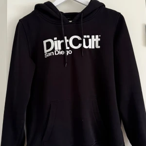 Svart hoodie från DirtCült - Säljer en svart hoodie från DirtCült med texten 'San Diego' tryckt på framsidan. Den har en klassisk design med huva och dragsnören samt en praktisk magficka. Perfekt för en avslappnad stil.