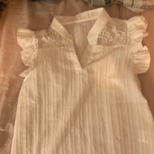 Vit blus med spetsdetaljer - Säljer en söt vit blus med spetsdetaljer vid axlarna och volangärmar. Blusen har en lätt v-ringning och är perfekt för en somrig look. Den är gjord i ett lätt och luftigt material. Dock en liten fläck på framsidan.