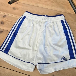 Vita sportshorts från Adidas - Säljer ett par vita sportshorts från Adidas med blåa detaljer och klassiska ränder på sidorna. Shortsen har en elastisk midja och är perfekta för träning eller fotboll. Adidas-loggan är broderad på benet.