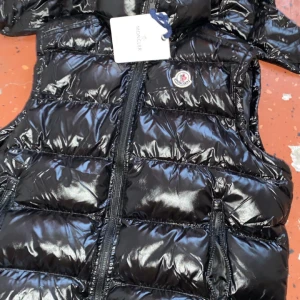 Svart dunväst från Moncler - Snygg svart dunväst från Moncler med glansig finish och dragkedja framtill. Perfekt för kyligare dagar när du vill hålla stilen. Moncler-logga på bröstet ger en exklusiv touch.
