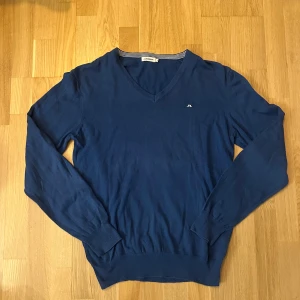 Blå v-ringad tröja J.Lindeberg - Klassisk blå v-ringad tröja från J.Lindeberg med diskret logga på bröstet. Långärmad modell i mjuk bomull, perfekt för lager-på-lager. Ribbstickade muddar och nederkant ger en clean look. Snygg till både jeans och chinos. Smått nopprig 