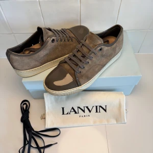 Lanvin skor  - Snygga bruna/grå sneakers från Lanvin med ovandel i mocka och glansig, lackad tå i mörkbrunt. Skorna har snörning och vit platt sula med diskret logga undertill. Perfekta för dig som gillar stilrena detaljer och exklusiv känsla. Obs igen og tillkommer vid köp!! Bara att höra av sig till oss vid minsta fundering😁