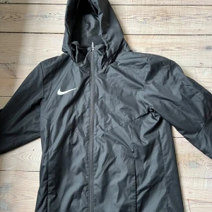 Svart Nike vindjacka med huva S - Helt ny svart vindjacka från Nike med huva och dragkedja framtill. Jackan har en stilren look med Nike-logga på bröstet och är tillverkad i lätt polyester. Perfekt för blåsiga dagar och enkel att matcha med streetwear eller träningskläder. PRISET ÄR INTE HUGGET I STEN