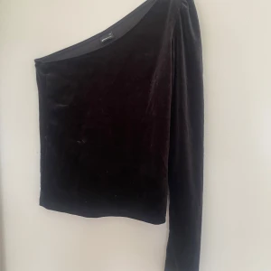 Svart fest topp - Säljer en svart sammets topp från Gina Tricot i storlek XS. Toppen har en snygg oneshoulder-design med lång ärm på ena sidan och ärmlös på andra. Mjuk och lyxig känsla, perfekt för att sticka ut lite extra.
