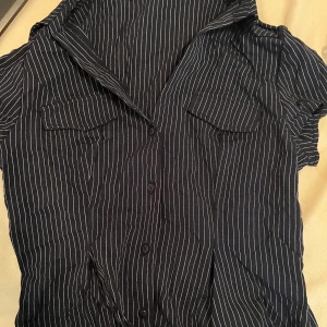 Svartvit randig skjorta med kort ärm - Snygg svart skjorta med vita ränder och korta ärmar. Skjortan har knappar framtill och två bröstfickor som model. Perfekt för dig som gillar en stilren och avslappnad look. Materialet känns lätt och passar bra till varmare dagar.