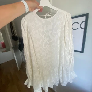 Vit blommig kortklänning med öppen rygg - Superfin vit kortklänning med blommigt mönster och volangdetaljer. Klänningen har långa ärmar, öppen rygg med knyt och en flowy passform. Perfekt för sommarens alla tillfällen när du vill känna dig extra söt och trendig.