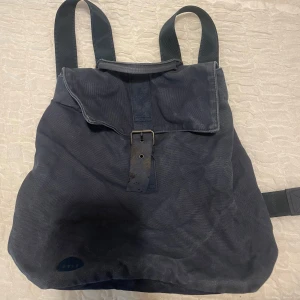Mörkblå ryggsäck från mi - En stilren mörkblå ryggsäck från mi med klassisk design. Väskan är tillverkad i kraftig bomullscanvas och har breda axelband samt ett lock som stängs med ett läderband och metallspänne. Perfekt för skolan!
