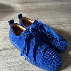 Blå mocka sneakers från Louboutin - Snygga blå sneakers i mocka från Christian Louboutin med coola nitar på tån och klassisk röd sula. Skorna har snörning och en låg profil, perfekt för dig som vill sticka ut med en unik detalj. Allt går i matchande blått, från sula till skosnören. Skorna är storlek 41 men passar upp till 43 skulle jag säga. Skicket är nästan sprillans nytt med en väldigt fräsch röd sula under.