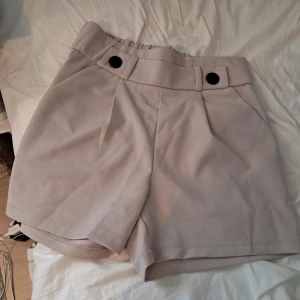 Beige shorts med knappar ONLY - Snygga beige shorts med hög midja och två svarta knappar framtill. De har en lös passform och är sydda i ett mjukt, vävt material som känns bekvämt mot huden. Perfekta för varma dagar och ger en clean och stilren look.