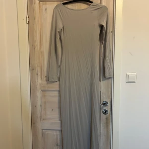 Grå långklänning från NA-KD - Snygg beige långklänning från NA-KD med lång ärm och en djup, öppen rygg. Klänningen är figurnära och har en enkel, stilren design i mjukt material. Perfekt för dig som gillar minimalistisk stil och vill sticka ut med en elegant siluett.