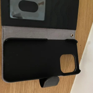 Svart iPhone skal, Ideal of Sweden - Snyggt svart plånboksfodral från Ideal of Sweden med plats för kort och ID-fönster. Fodralet är gjort i skinnimitation och har magnetstängning. Perfekt för dig som vill ha både mobil och kort på samma ställe. Enkel och stilren design. Denna är också helt oanvänd då jag ångrade mig direkt efter köpet.