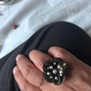 Blommig ring med pärlor  - Unik ring i antikguld med stor blomformad detalj. Blomman är dekorerad med vita pärlor och små gnistrande stenar som ger en cool vintagekänsla. Perfekt statement piece för dig som gillar att sticka ut med accessoarerna.