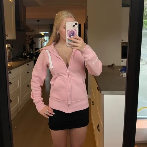 Rosa ziphoodie från Russemearch - Säljer en ljusrosa hoodie från Russemearch med dragkedja, huva och vita snören. Hoodien har broderad logga på ärmen och två fickor framtill. Mjuk och skön i bomullsmix, perfekt för chill dagar.