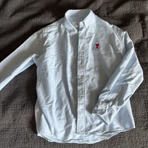 Randig skjorta från Ami Alexandre Mattiussi - Snygg ljusblå och vit randig skjorta från Ami Alexandre Mattiussi med klassisk krage och knäppning framtill. Skjortan har lång ärm och ett rött broderat hjärta med bokstaven A på bröstet. Tillverkad i bomull och passar perfekt för en clean och stilren look.