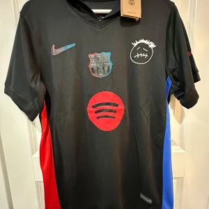 FC Barcelona Nike svart fotbollströja - Svart FC Barcelona fotbollströja från Nike med klubbmärke, Nike-logga och Spotify-logga på bröstet. Unikt tryck med vit smiley och röda/blå detaljer på sidorna. Tillverkad i lätt, ventilerande material som passar perfekt för match eller träning.